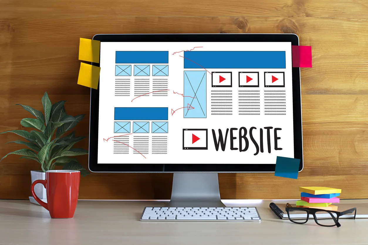 Widescreen monitor showing a website layout being rebuilt alongside design notes on a desk.