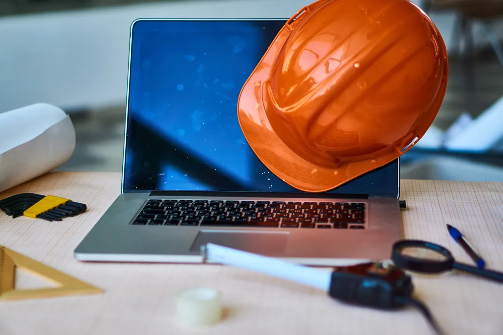 Site office desk with a laptop, printed construction drawings, and a hard hat